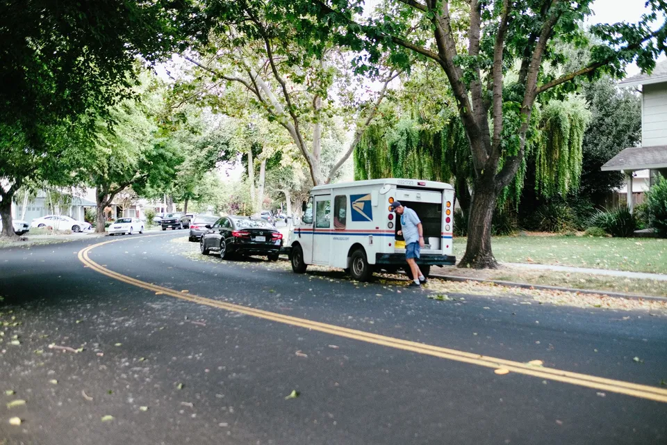 A US mail carrier retrieves packages from a postal van on a peaceful tree-lined street.