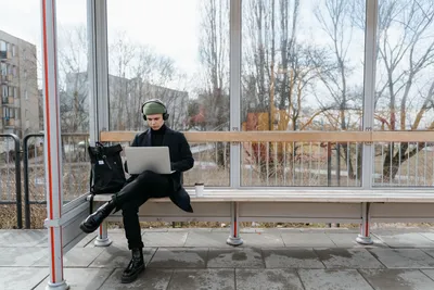 A young man works remotely on a laptop while sitting at a bus stop, showcasing modern digital nomad life.