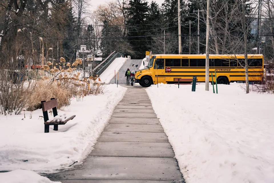 A school bus parked near a snowy sidewalk during winter, with people walking nearby.