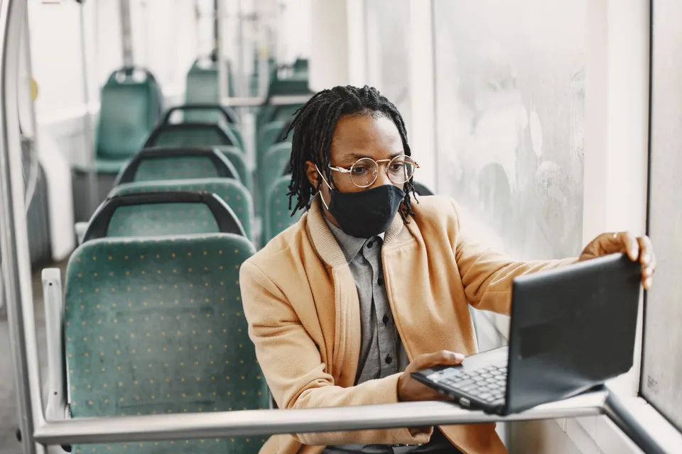 Young man in mask working on a laptop while commuting on a bus, showcasing safety and technology.