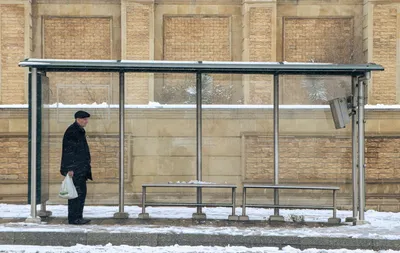 A man waits at a bus stop during a snowy winter day, holding a bag.